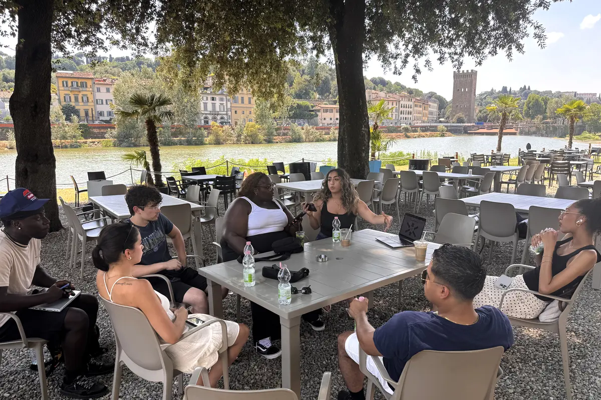 A young woman gives a presentation to six classmates while sitting outside under trees along a river in Italy.