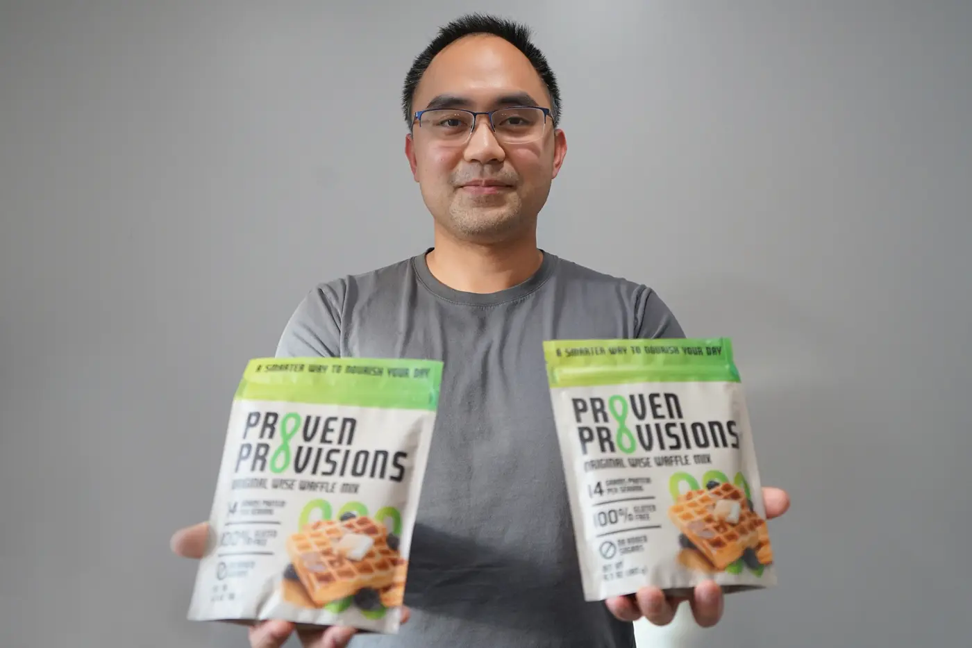 A man in glasses and a gray T-shirt holds two bags of waffle mix