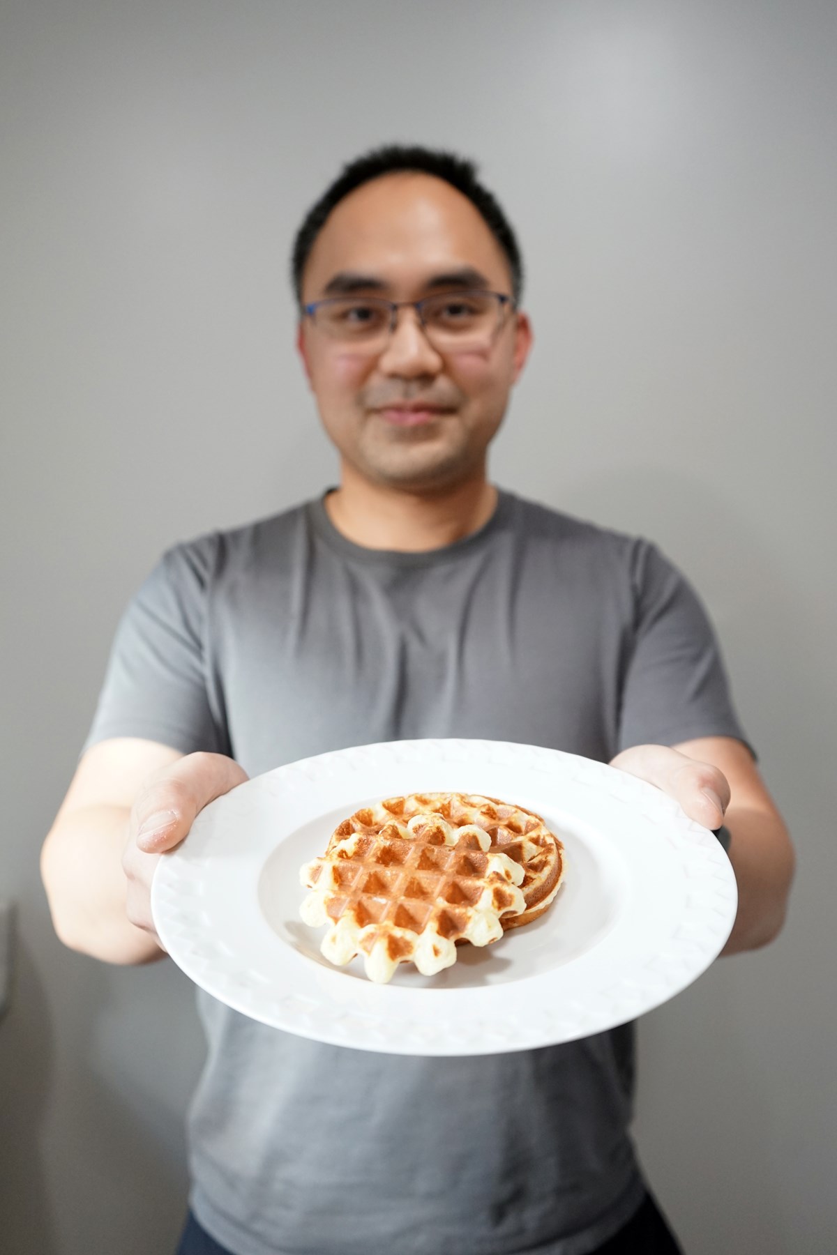 A man in glasses and a grey T-shirt holds a plate with two waffles.