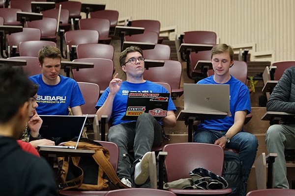 Three students in blue T-shirts work on laptops in a college auditorium