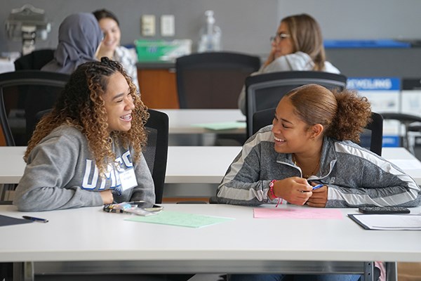 Two young women smile while talking to each other at a table in a conference room