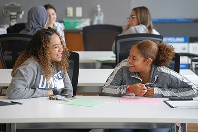 Two young women smile while talking to each other at a table in a conference room