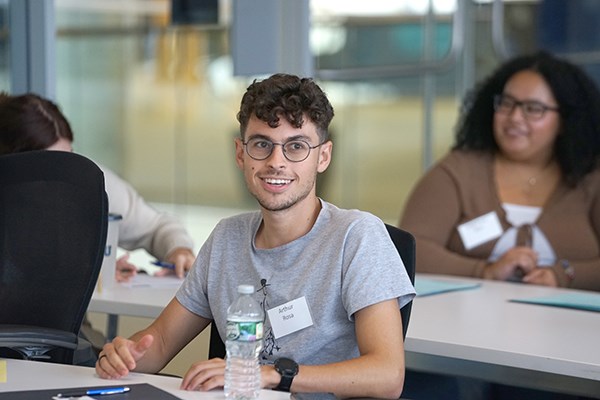 A young man in glasses smiles while listening to someone talk in a conference room