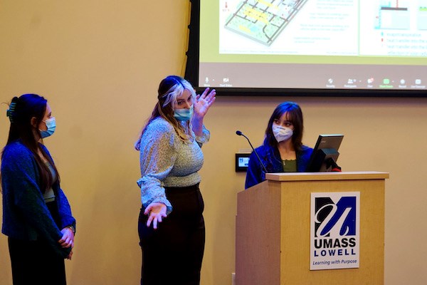 A student gestures with her hands while making a presentation with two other students at a podium