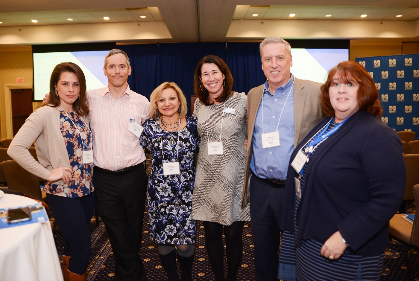 6 people standing together pose for a photo at the 2019 Annual Faculty Symposium with a blue UMass Lowell backdrop and others behind them.
