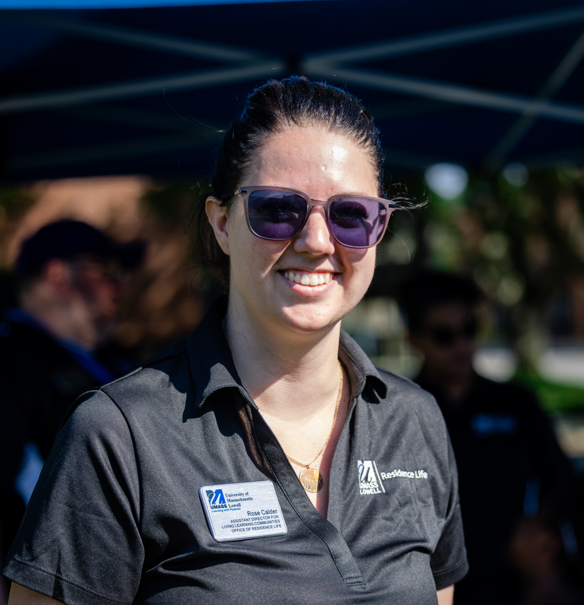 A woman smiles at the camera. Her black polo has the UMass Lowell logo and text "UMass Lowell".