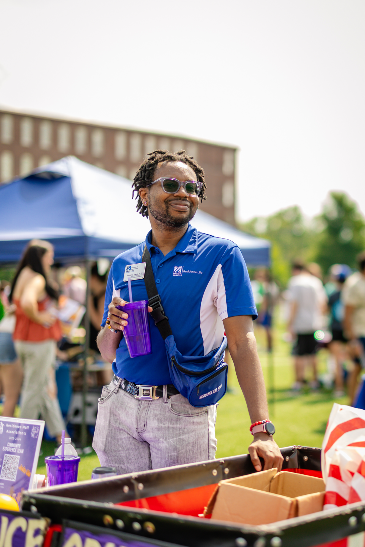 Staff standing, smiling holding a purple cup & wearing a blue UMass Lowell Residence Life shirt & Residence hall Association sling bag.