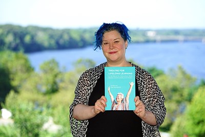 A woman poses for a photo holding a book while standing in front of a river