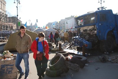 A young boy in a red coat and a man walk past a pile of tires and a broken down truck in a public square