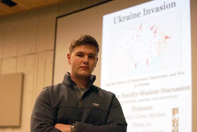 A student stands with his arms folded in front of a project screen that says "Ukraine Invasion"