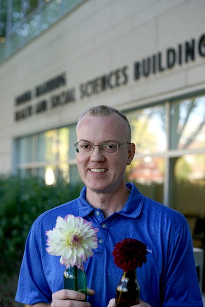 A man in a blue shirt smiles as he poses for a photo outside a building while holding flowers