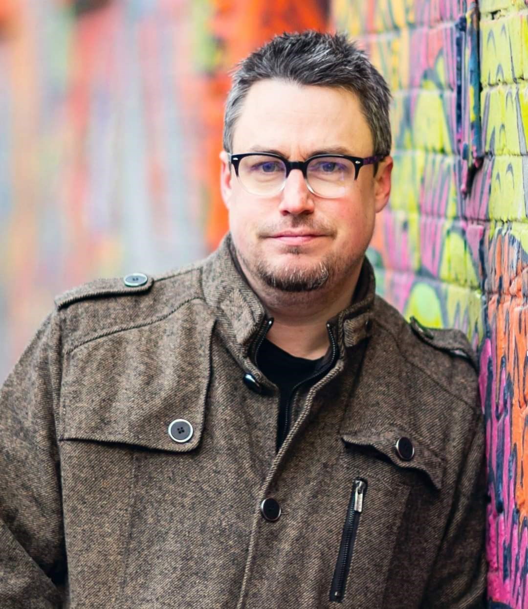 Ryan Cowley wearing glasses and a coat leaning against a multicolored/graffitied brick wall.