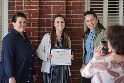 A woman holds a certificate while posing for a photo with two other women in front of a brick wall