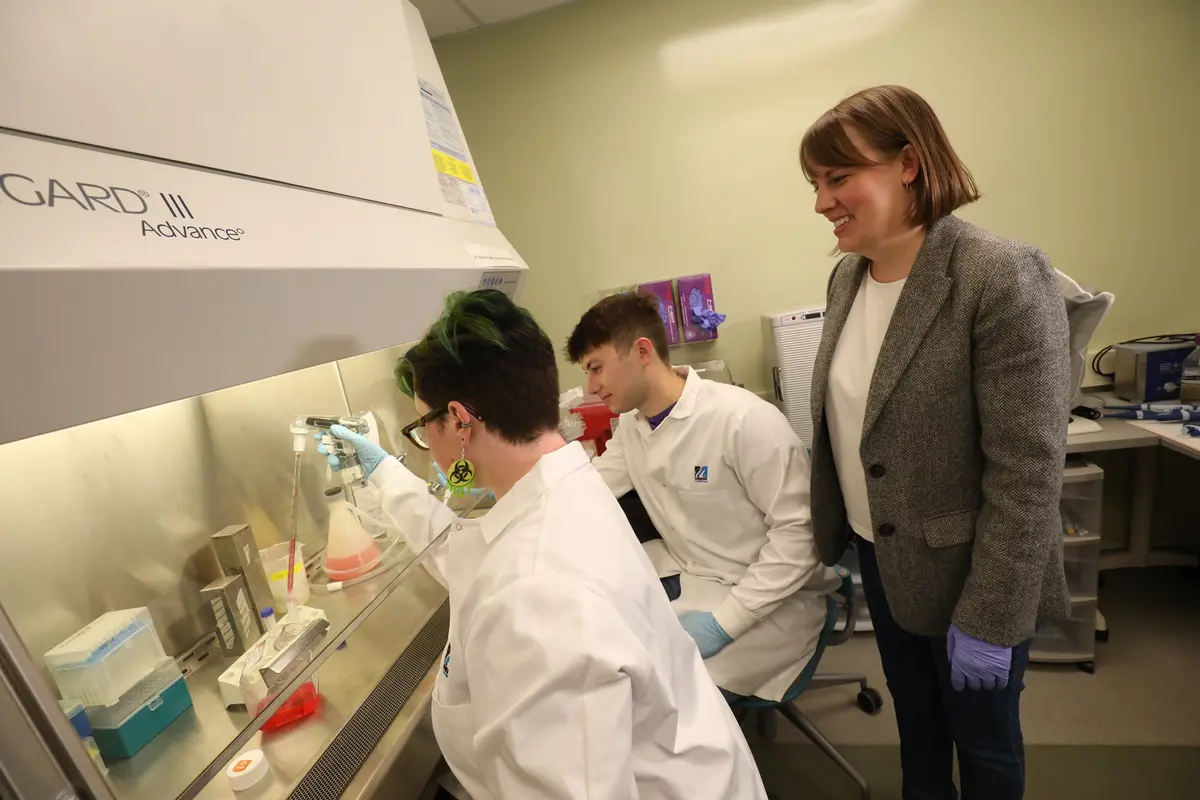 2 students in lab coats at GARD III Advance desk with woman standing behind them
