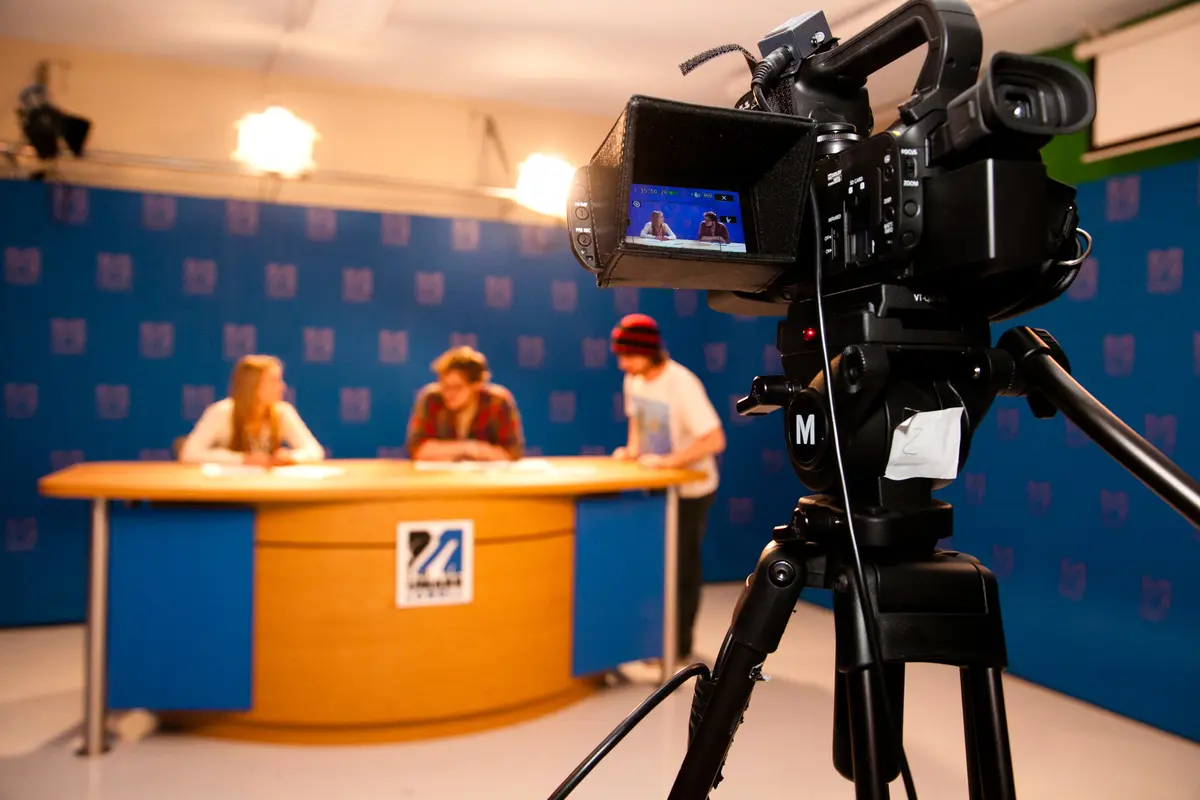 TV camera in foreground, 3 students blurry at anchor desk in studio with UMass Lowell logo background