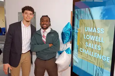 Two students stand next to a screen saying, "UMass Lowell Sales Challenge."