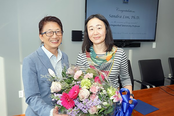 Two women pose for a photo in a conference room with a bouquet of flowers
