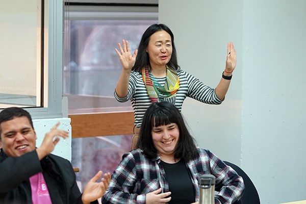A woman professor gestures with her hands while standing behind two smiling students in a classroom.