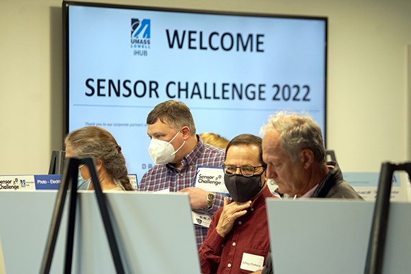 A woman and three men read poster boards in front of a monitor that says "Welcome Sensor Challenge 2022"