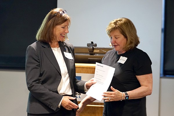 A smiling woman in a blazer presents a first-place certificate to another woman