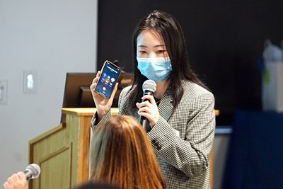 A woman in a face covering holds a microphone in one hand and an iphone in the other while looking at a person