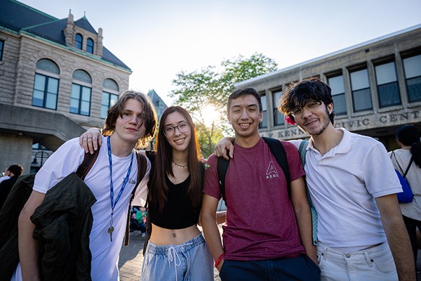 Four students pose for a photo outdoors while the sun shines through trees behind them