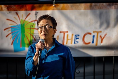 A woman wearing glasses holds a microphone and speaks outdoors