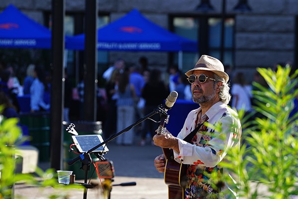 A man in sunglasses and hat plays guitar and sings into a mic outside