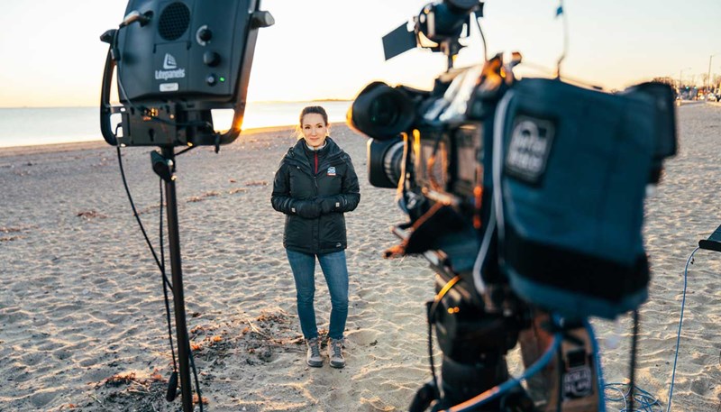UMass Lowell alumna and television meteorologist Shiri Spear stands on a beach in front of television cameras.