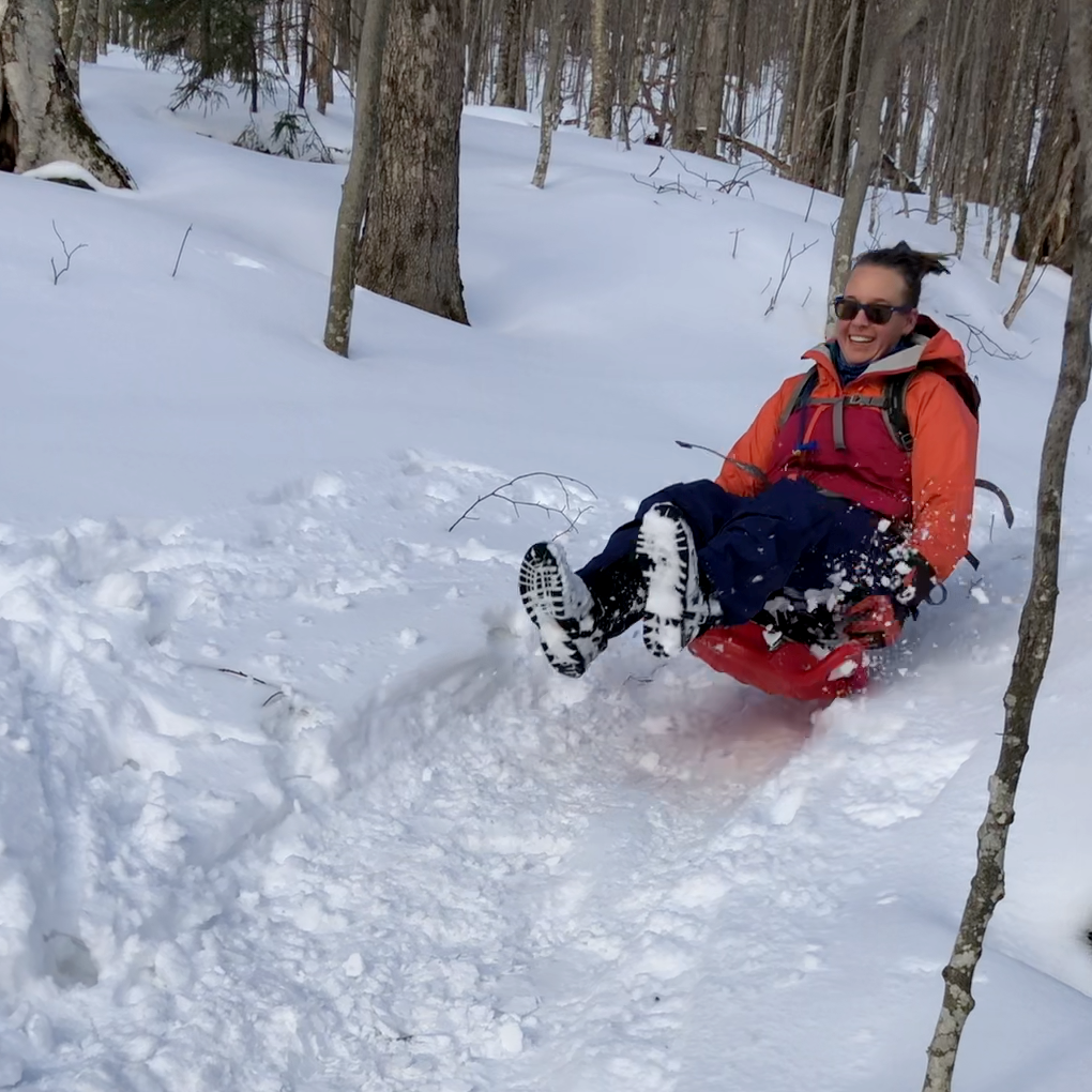 woman sleds down a trail in the woods with a big smile on her face