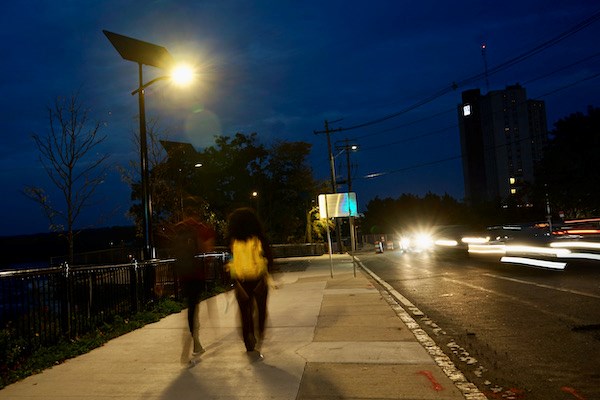 Two people walk down a sidewalk lit by solar street lights as car headlights blur past on their right