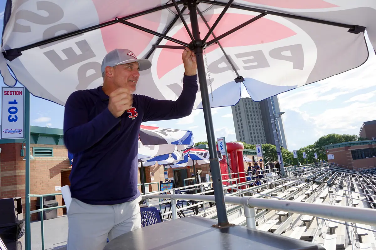 A man in a dark blue shirt and gray hat opens a patio umbrella on the concourse of a baseball stadium.