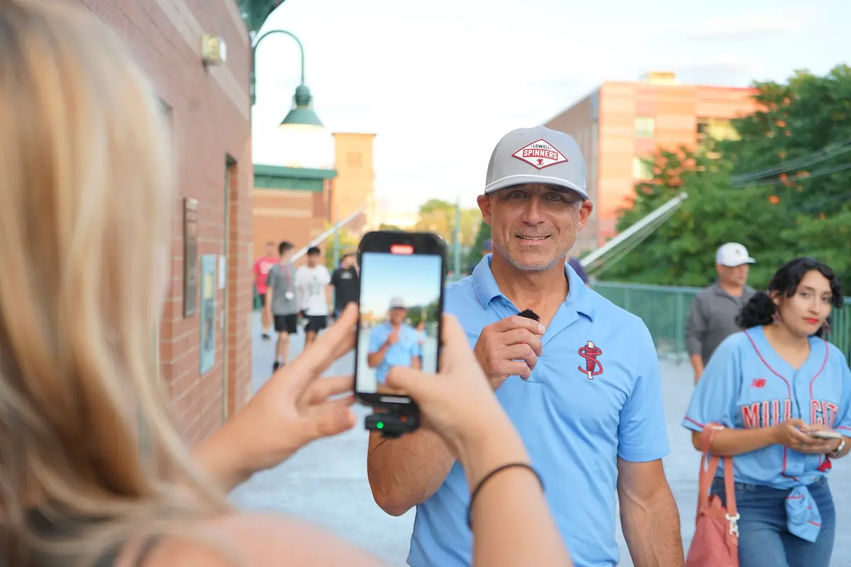 A man in a blue shirt and gray hat holds a microphone while being recorded by a woman with a smartphone.