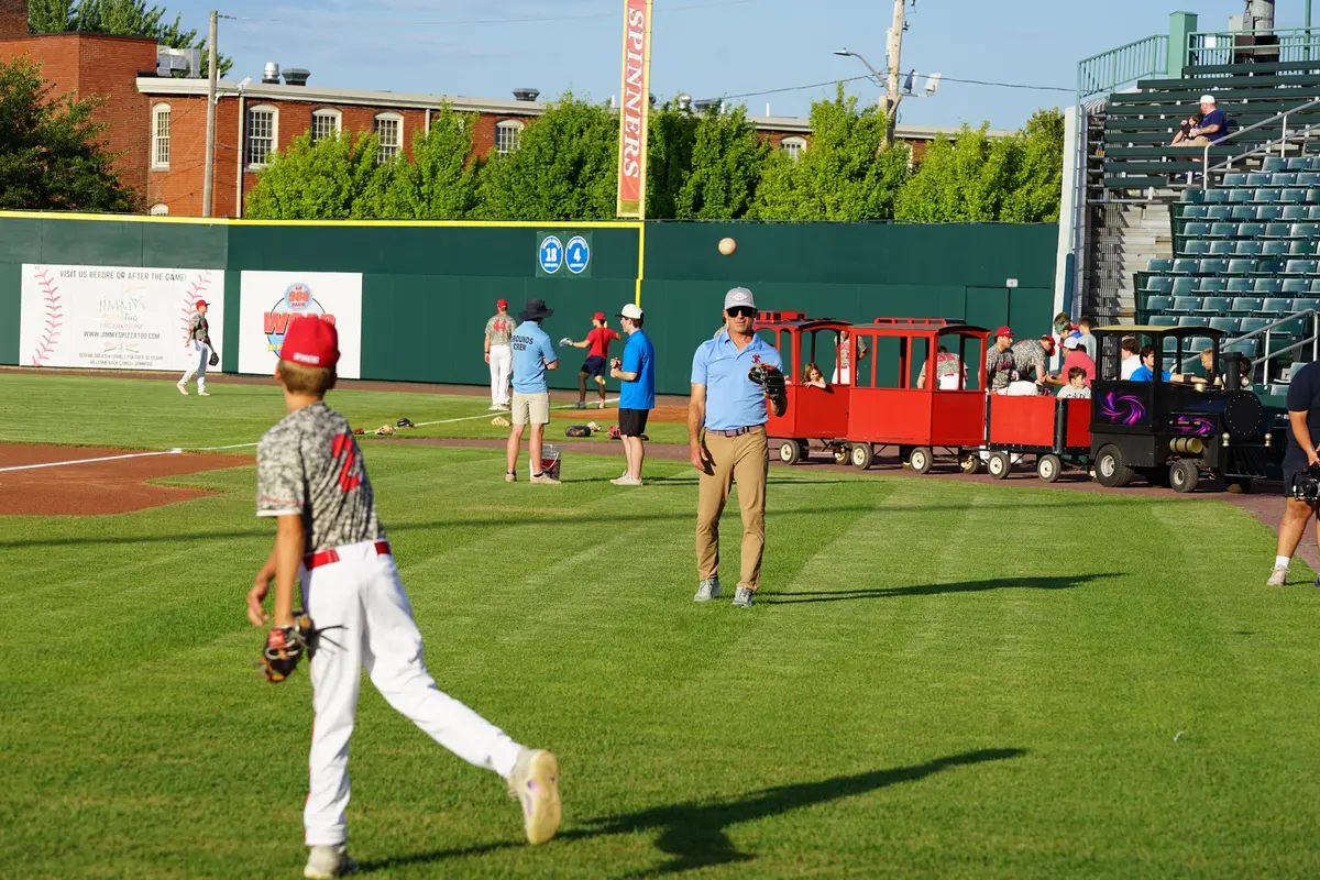 A man plays catch with his son on the field before a baseball game while a train ride passes behind him.