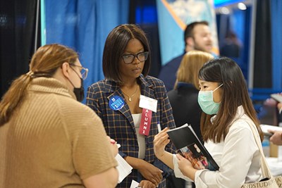Two women in glasses talk to a young woman wearing a mask at a career fair