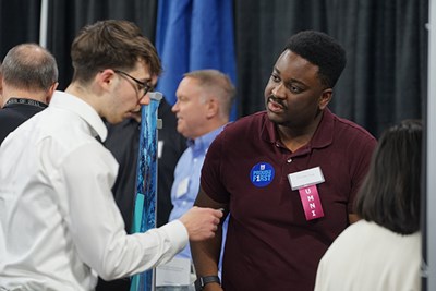A man in a maroon polo shirt listens as a young man in glasses talks to him at a career fair