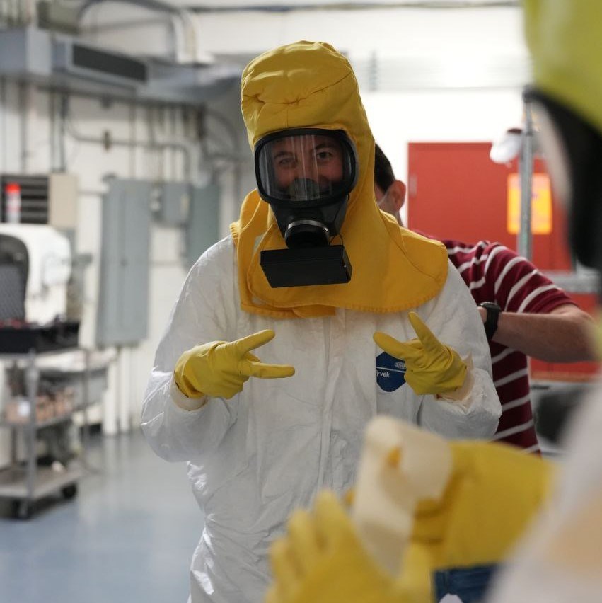 Simone Peironnet, Alumni, getting help donning radiation protection gear, respirator, suit, gloves, and coveralls while making two peace signs while another person in similar gear looks on.
