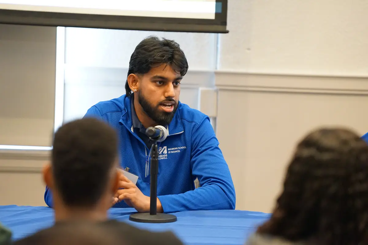 Srinivas Kagalkar sitting at a table with a blue tablecloth and speaking into a microphone to a group of students.