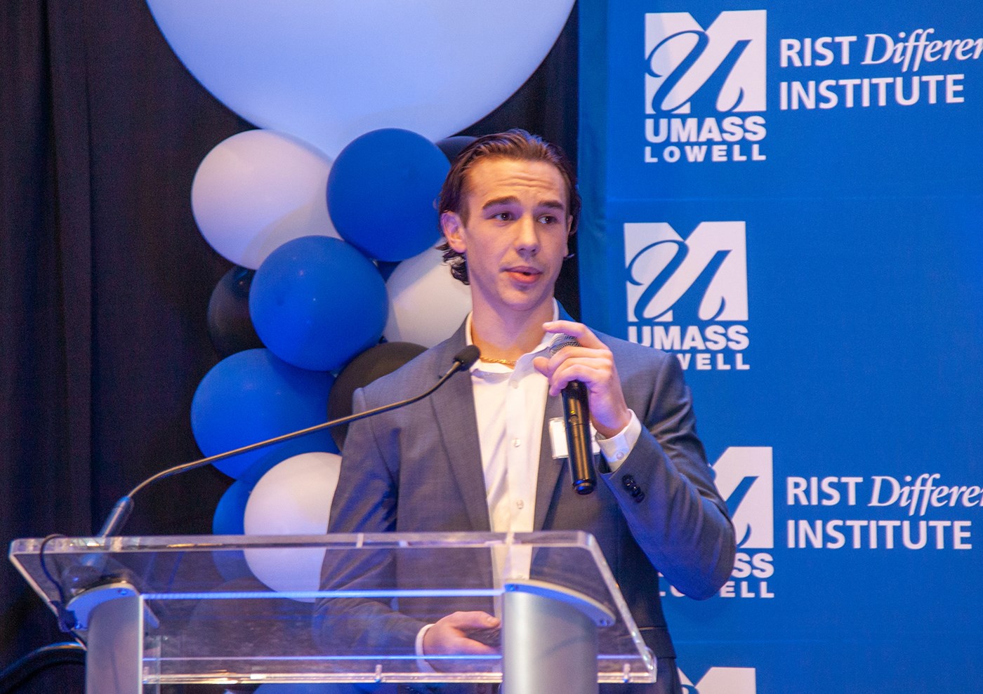 One of the members StockToMe team holding a microphone and speaking in front of blue UMass Lowell backdrop.