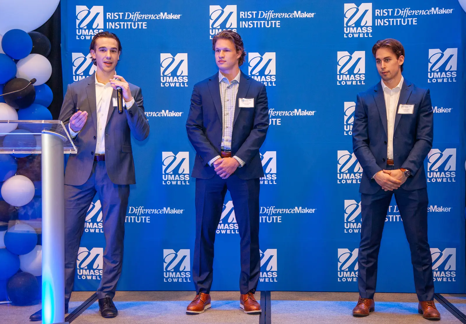 One of the members StockToMe team holding a microphone and speaking in front of blue UMass Lowell backdrop while two other members look on.