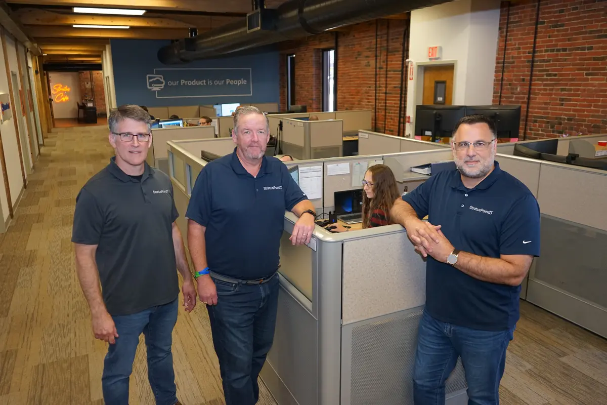 Three men in matching blue polo shirts stand next to a cubicle where a woman is working and pose for a photo.