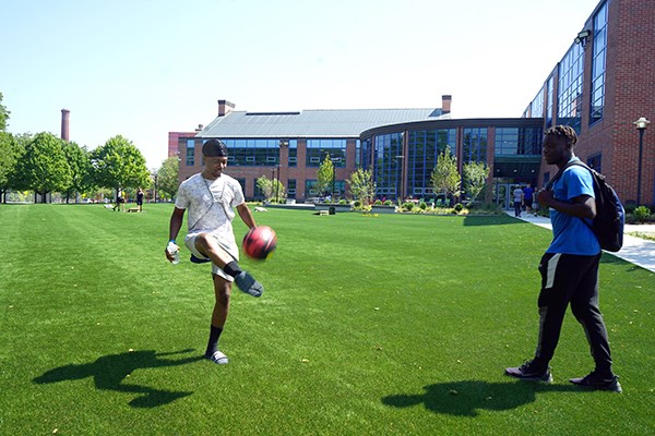 A man kicks a soccer ball on a turf field while another man looks on