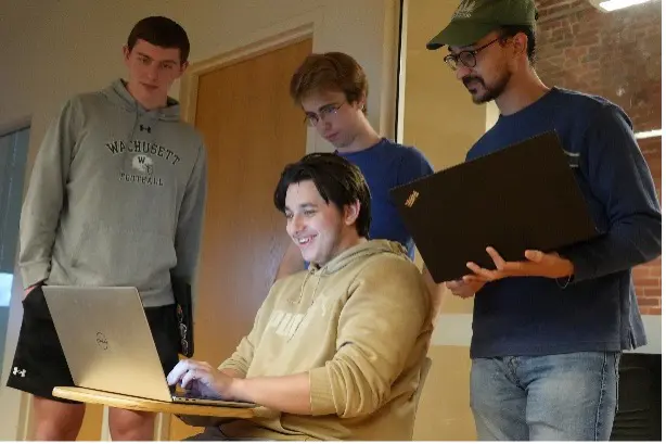 Three students stand behind and watch a fourth student computer on a computer.