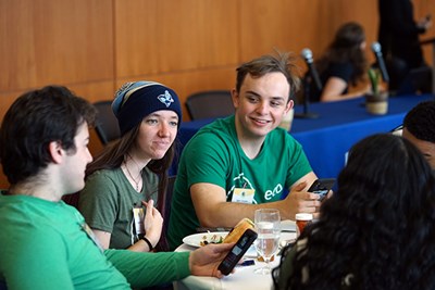 Three students wearing green shirts talk to each other while sitting at a table