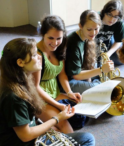 This one week, residential, musically intensive music camp is held mid-July each year for high school students grades 8-12. Students completing grade 12 are eligible pre-college and no audition is required. The four young ladies dressed in shorts and band camp t-shirts, two of them with french horns leaning against their legs, are seated on the floor conversing over an open piece of music. Selecting this link will take you to an information page for the group which will include how to register, cost to participate, and offer additional details.