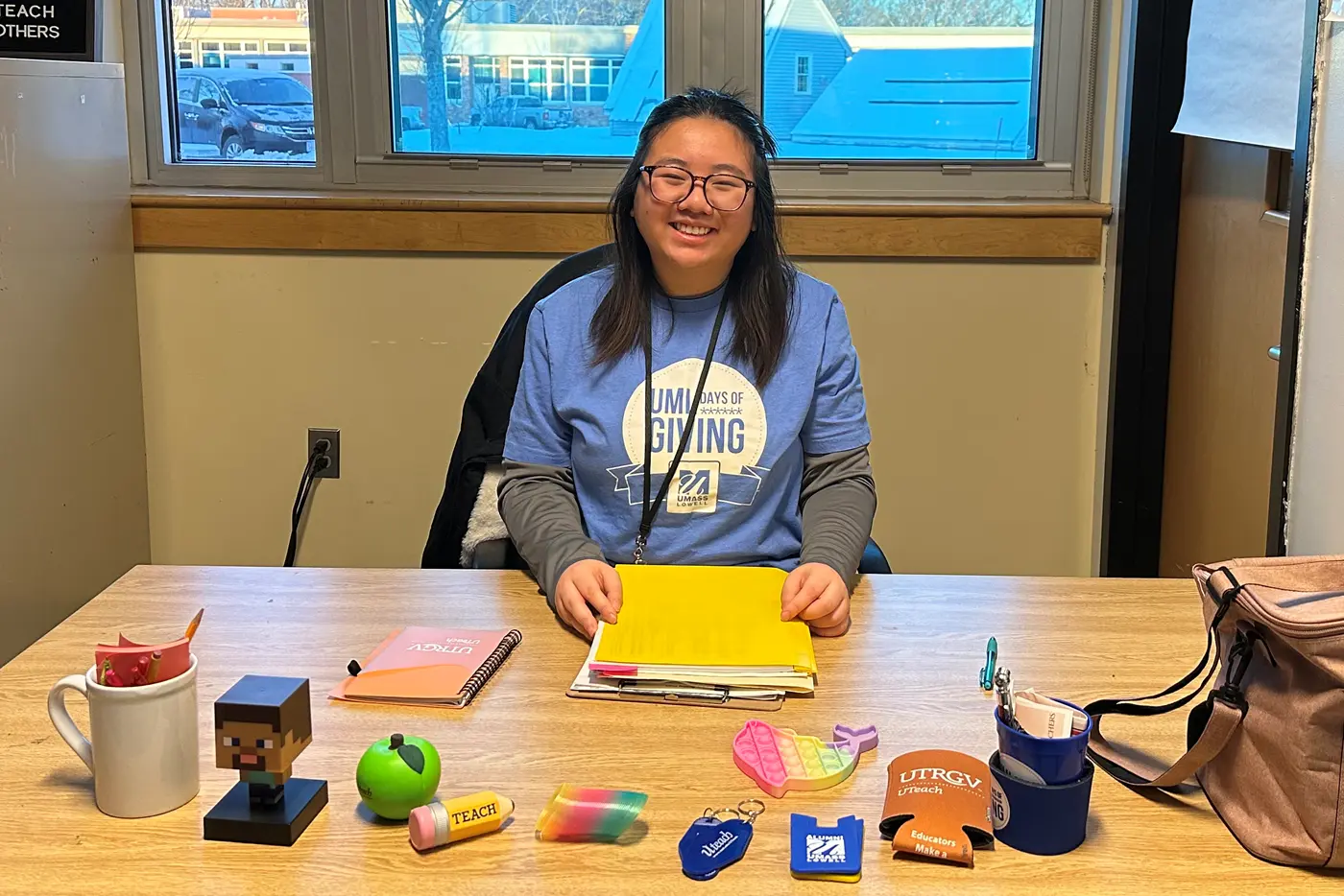 A young woman with dark hair and glasses poses for a photo while sitting at a desk, on which there are several giveaway items.