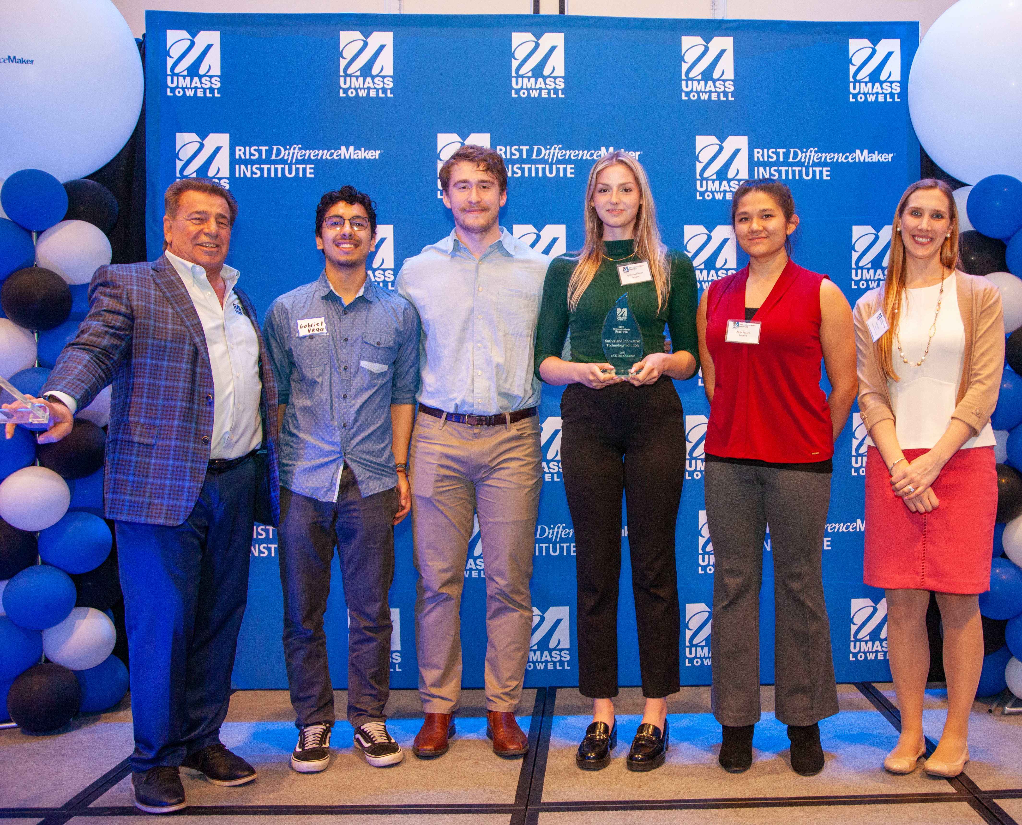 2 female and 2 male students from the Tendren team holding an award pose with Brian Rist and Holly Lalos of Difference Makers against a blue UMass Lowell backdrop.