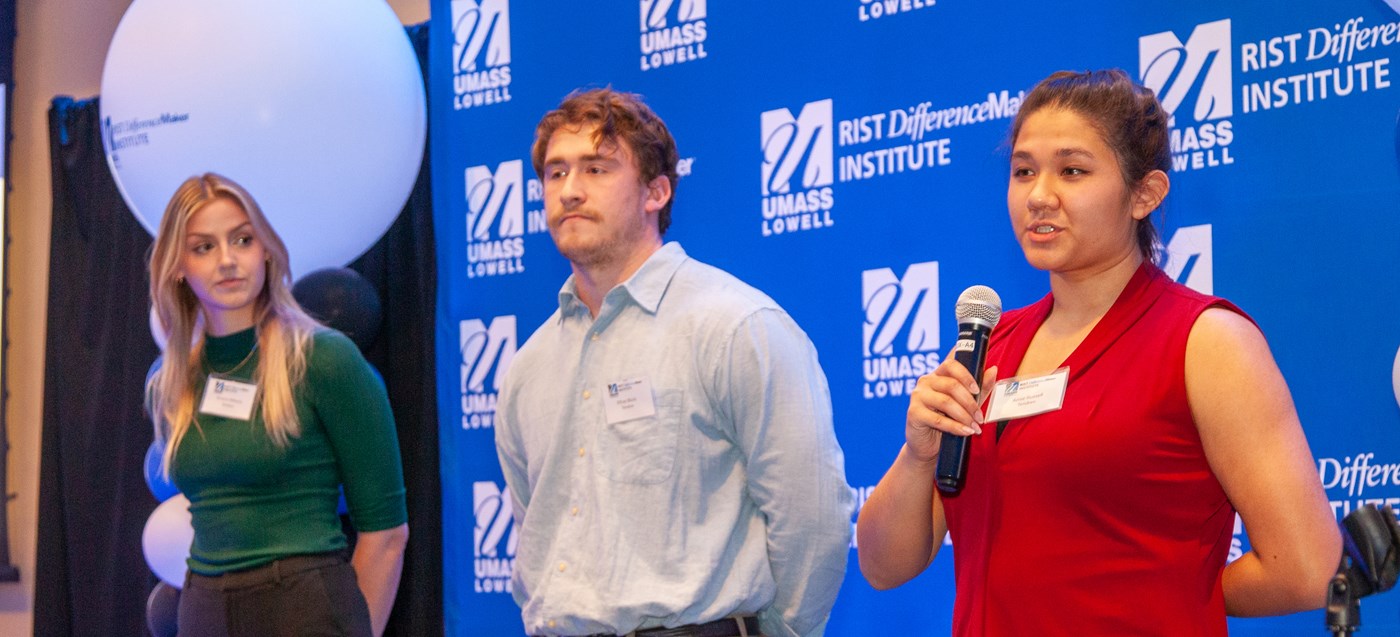 One of the members Tendren team holding a microphone and speaking in front of blue UMass Lowell backdrop as 2 other members look on.