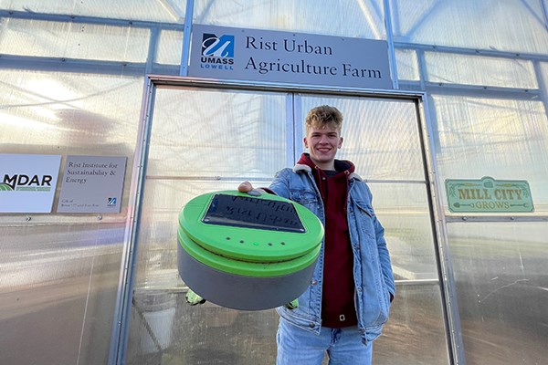 A male student holds a small green robot to the camera while standing in front of a greenhouse