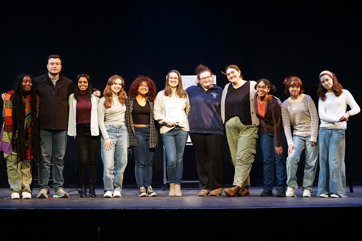 Eleven young people pose for a cast photo on a stage.